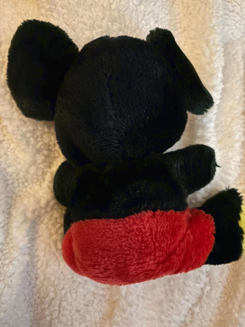 May include: A plush toy of a cartoon mouse, viewed from the back. The toy has black fur, large round ears, and red shorts. The toy's feet are yellow. The toy is sitting on a white, textured surface.