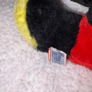 May include: Close-up of a plush toy featuring black, yellow, white, and red colors. The toy has a tag with text. The toy is on a white, textured surface.