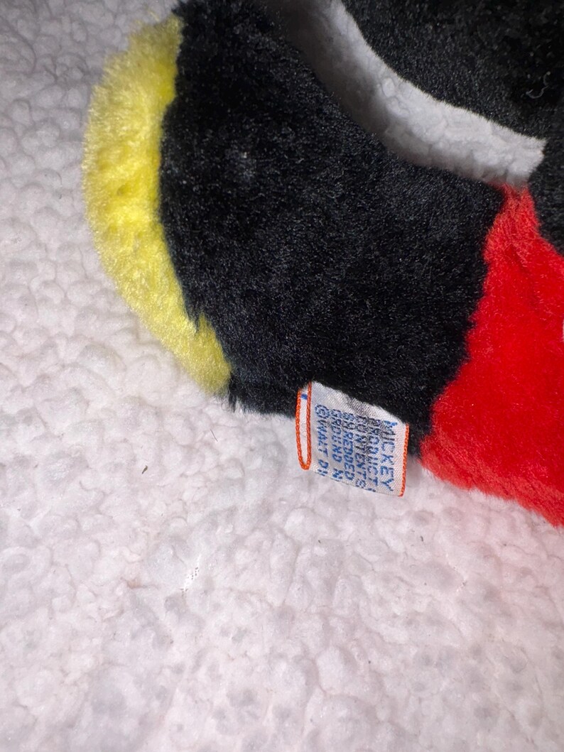 May include: Close-up of a plush toy featuring black, yellow, white, and red colors. The toy has a tag with text. The toy is on a white, textured surface.
