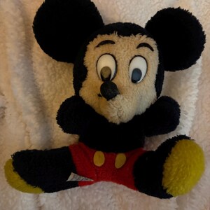 May include: A plush Mickey Mouse toy with black fur, large black ears, and a tan face. The toy wears red shorts with yellow buttons and has yellow soles. The toy is sitting on a white, fluffy surface.