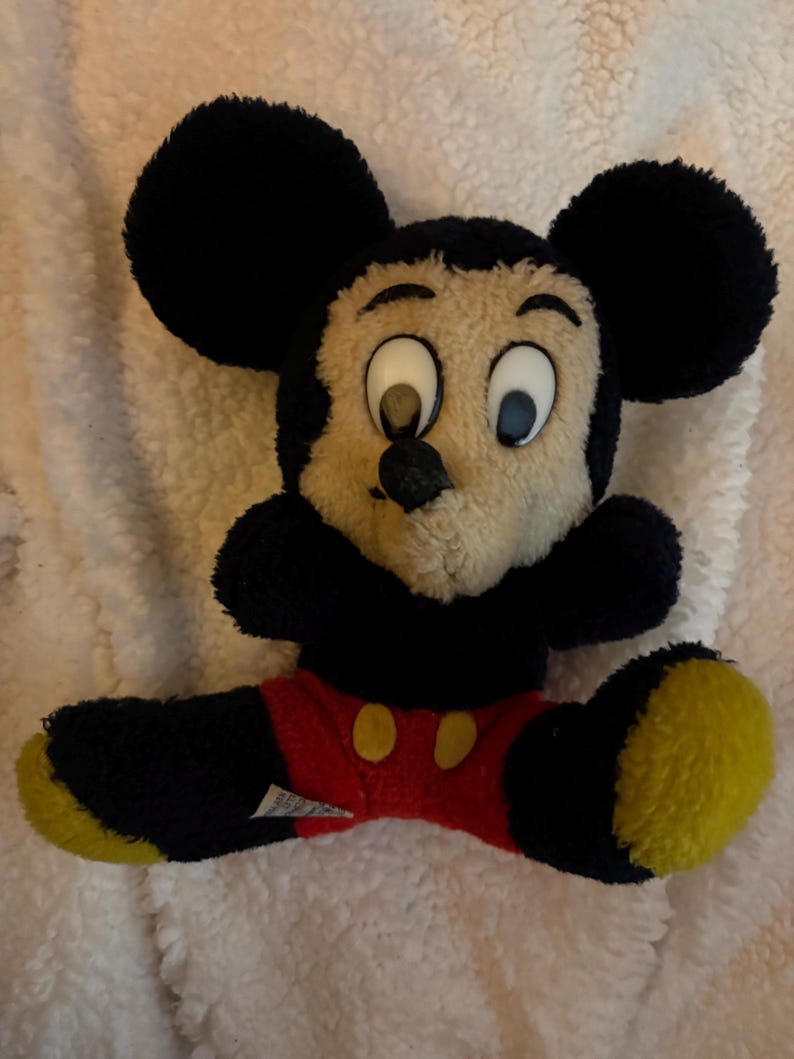 May include: A plush Mickey Mouse toy with black fur, large black ears, and a tan face. The toy wears red shorts with yellow buttons and has yellow soles. The toy is sitting on a white, fluffy surface.