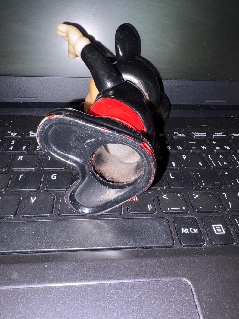 Pu&ograve; includere: Una statuetta in ceramica nera e rossa di Topolino in posizione eretta con il braccio destro alzato. La statuetta &egrave; su una tastiera di un computer portatile nero.