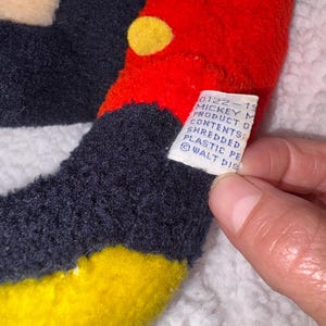 May include: Close-up of a vintage Mickey Mouse plush toy. The toy features a red head with yellow button-like accents, black ears, and a yellow and black body. A white tag with text is visible.
