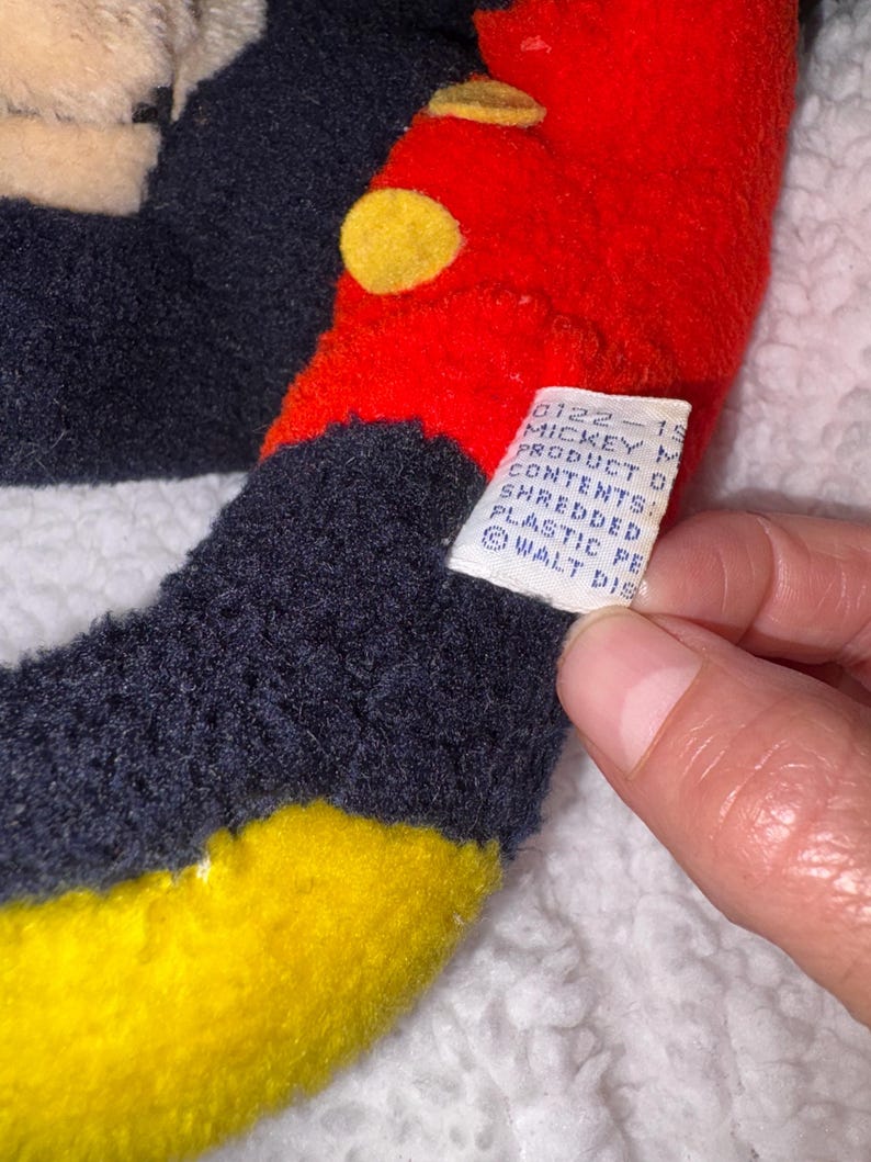 May include: Close-up of a vintage Mickey Mouse plush toy. The toy features a red head with yellow button-like accents, black ears, and a yellow and black body. A white tag with text is visible.