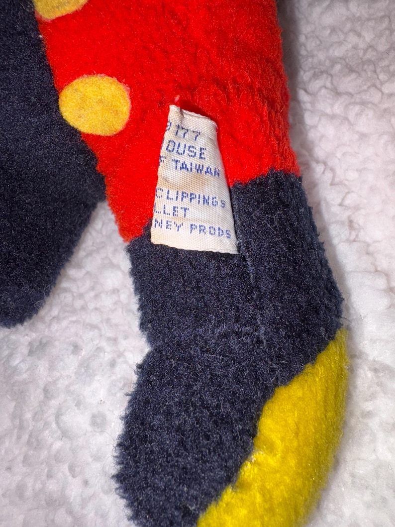 May include: Close-up of a plush toy with red, navy blue, and yellow sections. The red section has yellow circles. A tag reads "177 Duse Taiwan, Clippings Ballet, Disney Props."