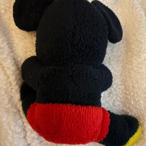 May include: A black plush toy of a cartoon mouse, viewed from the back. The toy has large, round ears, a black body, red shorts, and yellow feet. The toy is sitting on a white, textured surface.