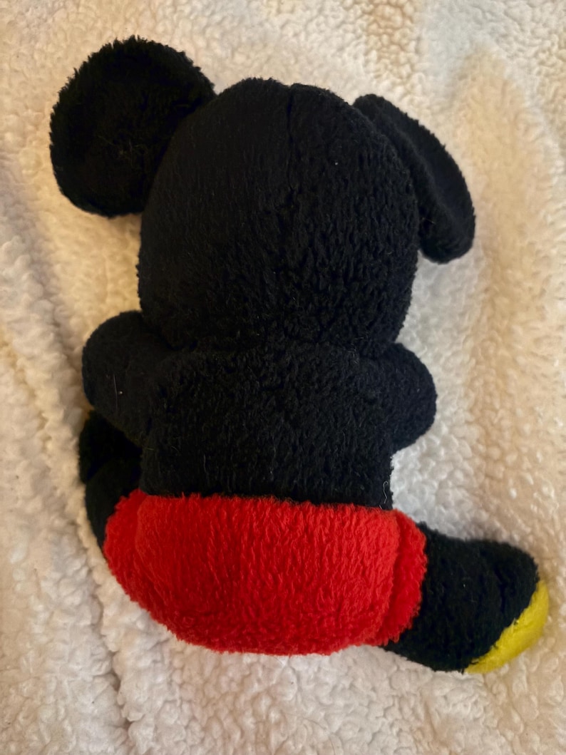 May include: A black plush toy of a cartoon mouse, viewed from the back. The toy has large, round ears, a black body, red shorts, and yellow feet. The toy is sitting on a white, textured surface.