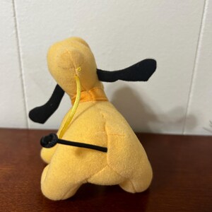 Pu&ograve; includere: Un cane di peluche giallo con orecchie nere e coda nera. Il cane indossa un collare giallo con una striscia arancione.