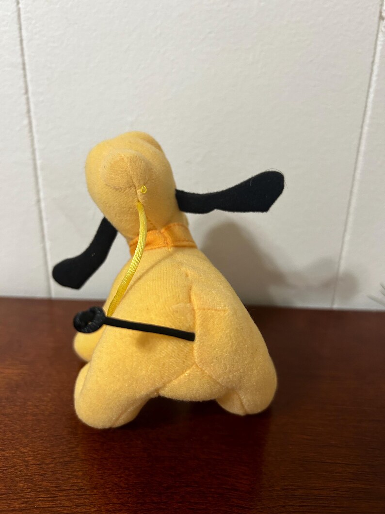 Pu&ograve; includere: Un cane di peluche giallo con orecchie nere e coda nera. Il cane indossa un collare giallo con una striscia arancione.