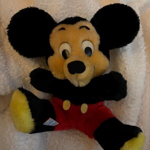 May include: A vintage Mickey Mouse plush toy. The plush features a black head with large round ears, a tan face, and black eyes. It wears red shorts with yellow buttons, black arms and legs, and yellow feet.