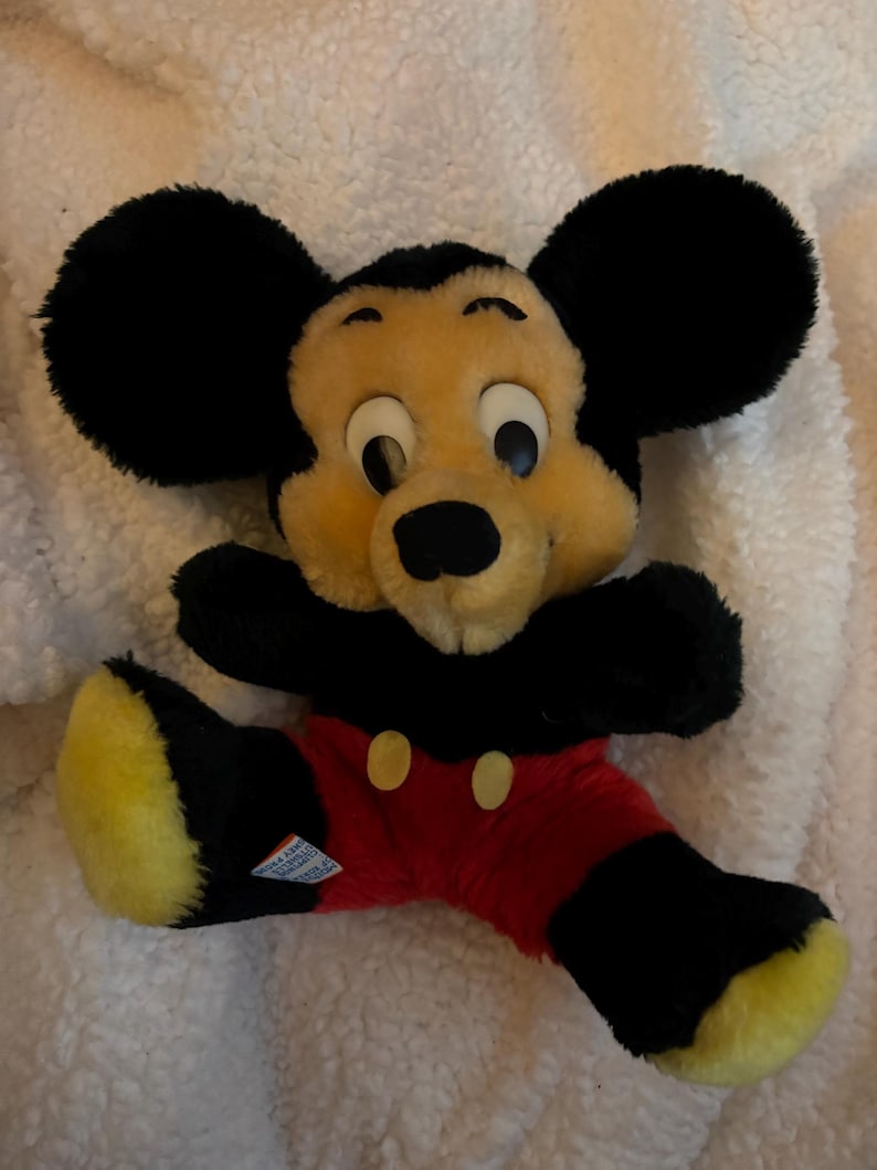 May include: A vintage Mickey Mouse plush toy. The plush features a black head with large round ears, a tan face, and black eyes. It wears red shorts with yellow buttons, black arms and legs, and yellow feet.