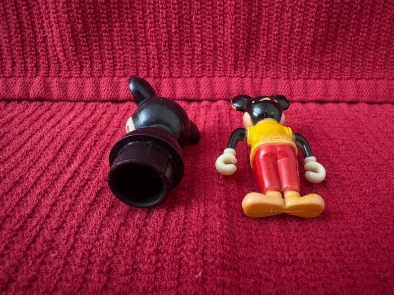 May include: Two vintage plastic Mickey Mouse figurines. One is black with a hat and the other is red, yellow, and black with a yellow shirt and red shorts.