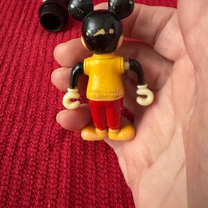 May include: Vintage plastic Mickey Mouse toy with a yellow shirt, red pants, and white gloves. The toy has a black head with white ears and a black nose. The toy is marked "Walt Disney Productions" on the back.