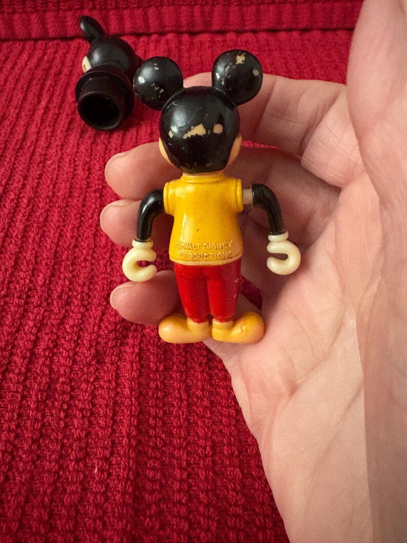 May include: Vintage plastic Mickey Mouse toy with a yellow shirt, red pants, and white gloves. The toy has a black head with white ears and a black nose. The toy is marked "Walt Disney Productions" on the back.