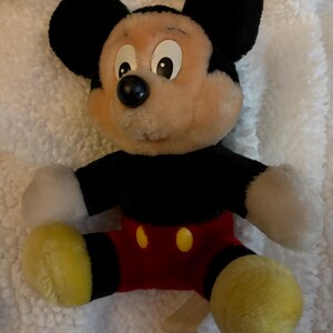 May include: A plush Mickey Mouse toy. The toy has a black body, ears, and a black nose. It has a tan face, white gloves, red shorts, and yellow shoes. The toy is sitting on a white, fluffy surface.