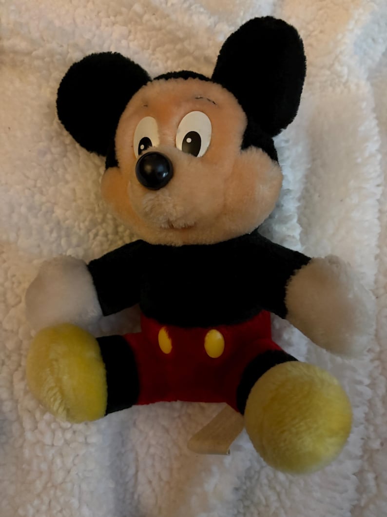 May include: A plush Mickey Mouse toy. The toy has a black body, ears, and a black nose. It has a tan face, white gloves, red shorts, and yellow shoes. The toy is sitting on a white, fluffy surface.
