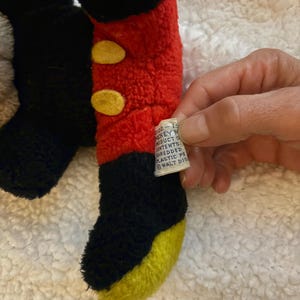 May include: Close-up of a plush toy leg, featuring black, red, and yellow colors. The leg has a red upper portion with two yellow circular accents, transitioning to black and then yellow at the foot. A tag is visible.