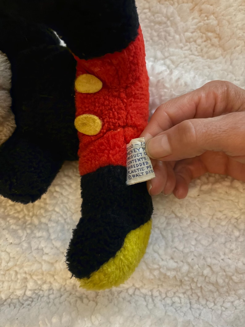 May include: Close-up of a plush toy leg, featuring black, red, and yellow colors. The leg has a red upper portion with two yellow circular accents, transitioning to black and then yellow at the foot. A tag is visible.