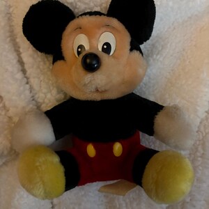 May include: A plush Mickey Mouse toy with a black body, ears, and a red pair of shorts. The toy has a cream-colored face, white gloves, and yellow feet. The toy is sitting on a white, fluffy surface.