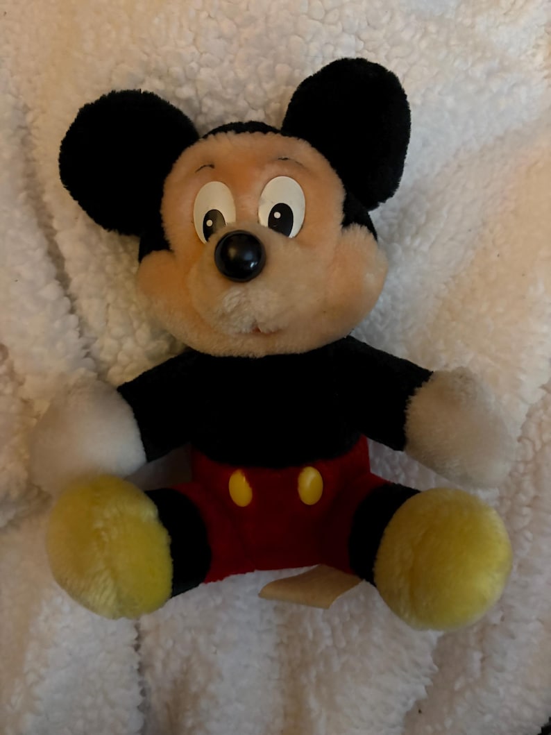 May include: A plush Mickey Mouse toy with a black body, ears, and a red pair of shorts. The toy has a cream-colored face, white gloves, and yellow feet. The toy is sitting on a white, fluffy surface.