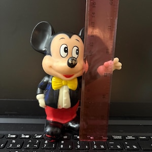 Pu&ograve; includere: Una statuetta vintage di Topolino con uno smoking nero, un papillon giallo e pantaloni rossi. La statuetta &egrave; alta circa 17 cm ed &egrave; realizzata in plastica.