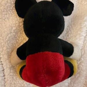 May include: A plush toy of a famous cartoon character, viewed from the back. The toy features black ears, a black head and body, red shorts, white hands, and yellow and black shoes. The toy is on a white, textured surface.