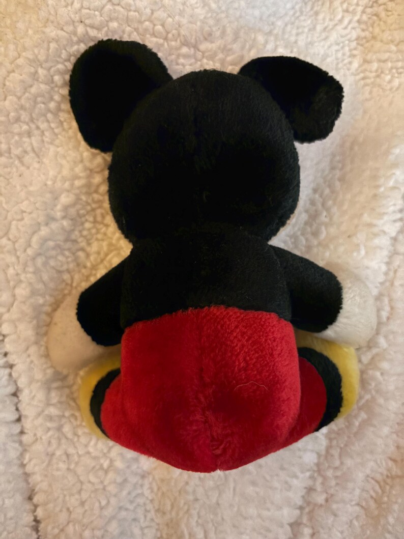 May include: A plush toy of a famous cartoon character, viewed from the back. The toy features black ears, a black head and body, red shorts, white hands, and yellow and black shoes. The toy is on a white, textured surface.