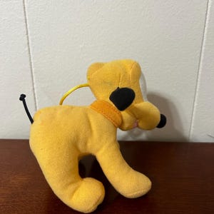 Pu&ograve; includere: Un cane di peluche giallo con orecchie nere e naso nero. Il cane indossa un collare giallo.
