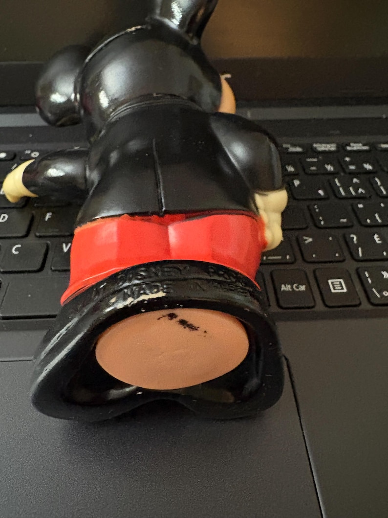 May include: Black and red plastic Mickey Mouse figurine with a black base. The figurine is wearing a black suit and red shorts. The base has the text "MADE IN HONG KONG" on it.