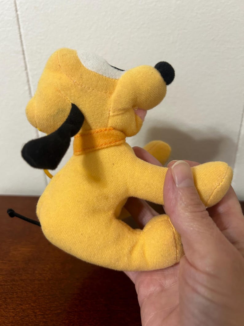 Pu&ograve; includere: Un cane di peluche giallo con orecchie nere e coda nera. Il cane ha una macchia bianca sul viso e indossa un collare arancione.