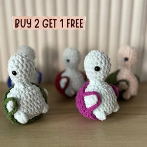 Può includere: Una collezione di figurine di tartarughe fatte a mano all'uncinetto in vari colori. Le tartarughe hanno corpi bianchi, occhi neri e gusci colorati in verde, rosa e rosso. Il testo "BUY 2 GET 1 FREE" è visualizzato sopra le tartarughe.