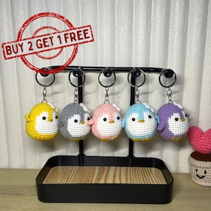 Puede incluir: Cinco llaveros de pingüino de ganchillo en amarillo, gris, rosa, azul y morado, cada uno con vientre blanco y ojos negros. Los llaveros cuelgan de un expositor de metal negro. El texto "BUY 2 GET 1 FREE" está en un círculo rojo.