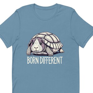 Op de afbeelding: Lichtblauw t-shirt met een afbeelding van een cavia met een schildpadschild en de witte tekst "BORN DIFFERENT". De cavia heeft een wit gezicht en een grijs lichaam. Het shirt is gemaakt van een zacht materiaal.