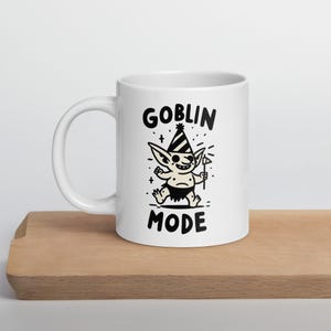 May include: White ceramic mug with the words "GOBLIN MODE" and a cartoon goblin illustration in black. The goblin is wearing a party hat and holding a small flag. The mug is sitting on a wooden surface.