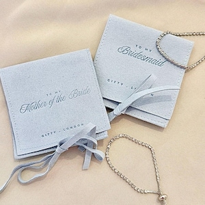 Puede incluir: Bolsas de terciopelo azul claro con las inscripciones "TO MY Mother of the Bride" y "TO MY Bridesmaid". También se ven dos pulseras de plata con piedras transparentes. Las bolsas están atadas con cintas a juego.