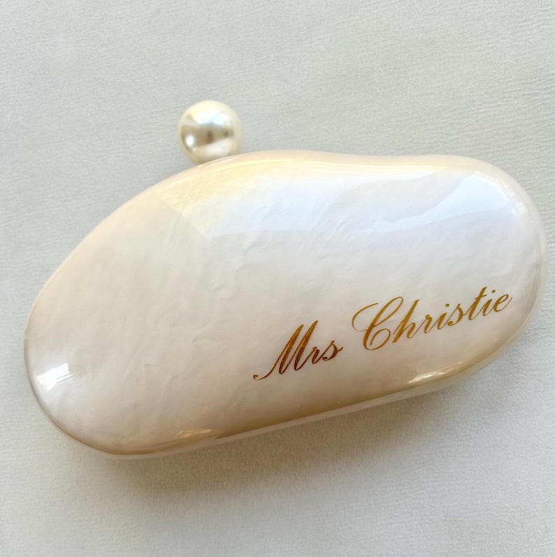 May include: A pearlescent white clutch with a rounded shape and a pearl accent. The words "Mrs Christie" are written in gold script on the front of the clutch. The clutch is a stylish accessory.