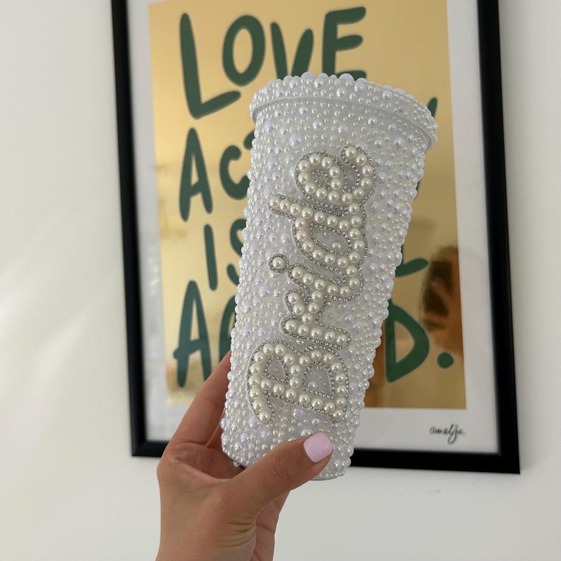 Op de afbeelding: Een witte beker bedekt met kleine, witte parels. Het woord "Bride" is geschreven met grotere parels en zilverkleurige kralen. De beker wordt vastgehouden voor een ingelijste poster met de woorden "LOVE ACTS IS ALL D." in groen.
