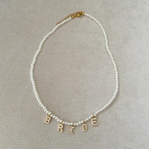 May include: A white pearl necklace with gold-colored letters spelling "BRIDE". The letters are encrusted with small, clear crystals. The necklace has a gold-colored clasp.