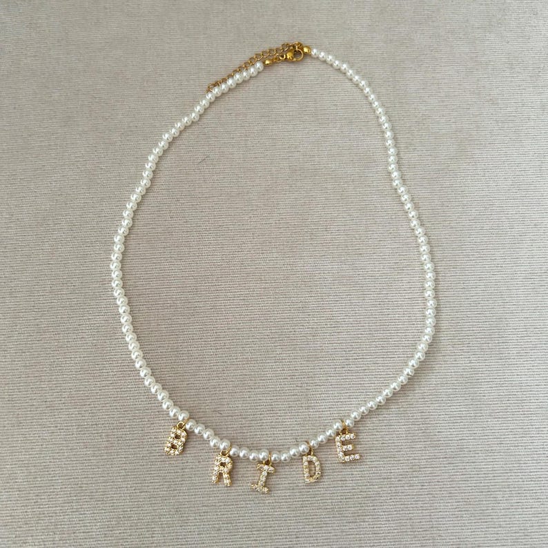 May include: A white pearl necklace with gold-colored letters spelling "BRIDE". The letters are encrusted with small, clear crystals. The necklace has a gold-colored clasp.