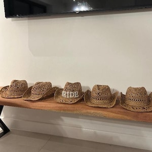Puede incluir: Cinco sombreros de vaquero de paja tejida con un dise&ntilde;o de tejido abierto. Un sombrero tiene la palabra "RIDE" en pedrer&iacute;a plateada. Los sombreros se exhiben sobre una superficie de madera.