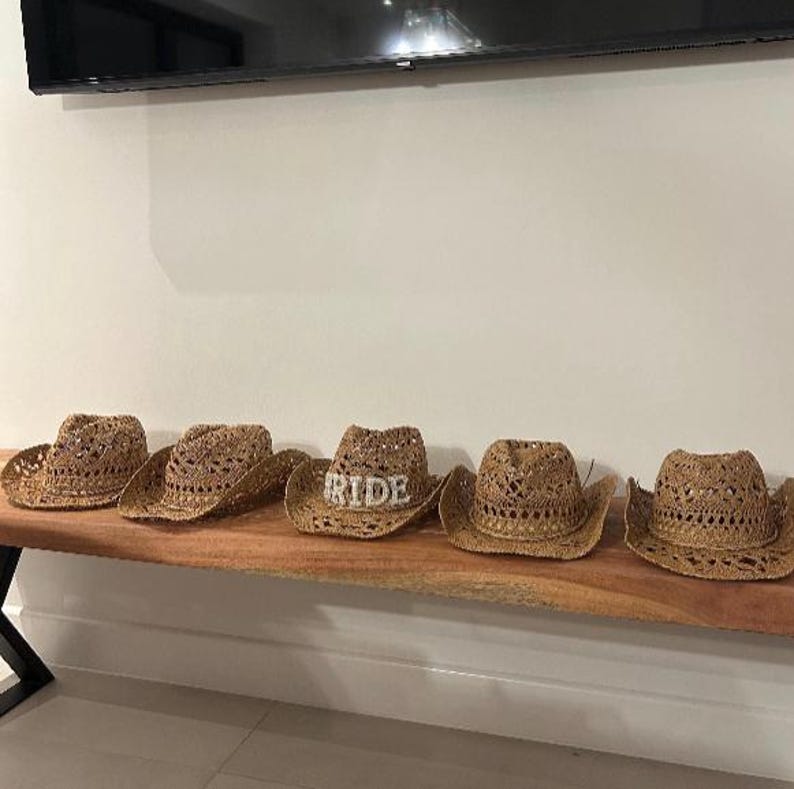 Puede incluir: Cinco sombreros de vaquero de paja tejida con un dise&ntilde;o de tejido abierto. Un sombrero tiene la palabra "RIDE" en pedrer&iacute;a plateada. Los sombreros se exhiben sobre una superficie de madera.