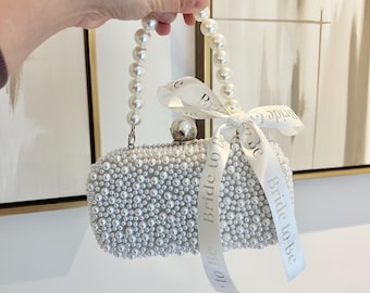 May include: A white, rectangular clutch covered in small, round pearls. The handle is made of larger pearls. A white ribbon tied around the top of the clutch reads "Bride to Be". The clutch has a silver clasp and a decorative silver ball on top.
