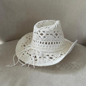 Puede incluir: Un sombrero de vaquero blanco de tejido abierto con un lazo decorativo de cuerda. El sombrero presenta un ala ancha y un patr&oacute;n tejido detallado. El sombrero est&aacute; hecho de un material de color claro.