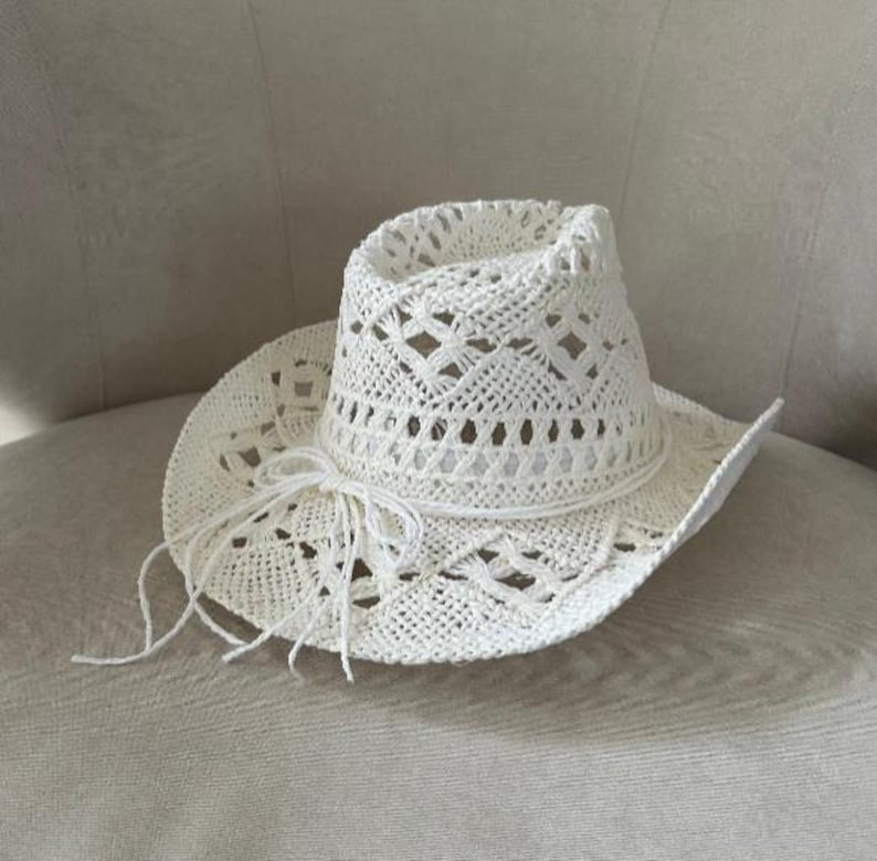 Puede incluir: Un sombrero de vaquero blanco de tejido abierto con un lazo decorativo de cuerda. El sombrero presenta un ala ancha y un patr&oacute;n tejido detallado. El sombrero est&aacute; hecho de un material de color claro.
