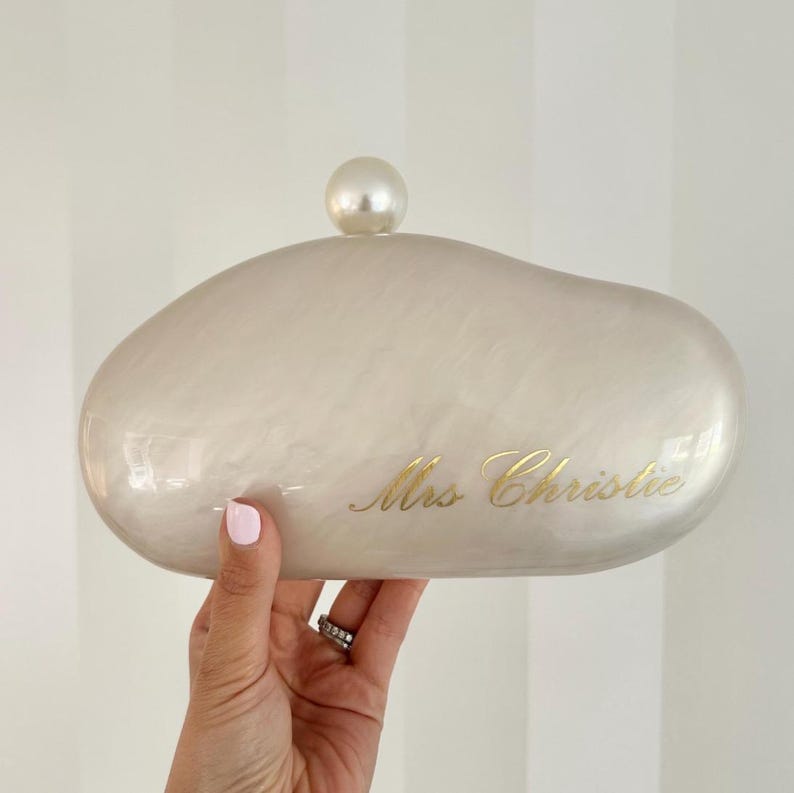 May include: A pearlescent clutch with a rounded shape and a pearl-like clasp. The text "Mrs Christie" is inscribed in gold script on the front. The clutch is held by a hand with a ring.