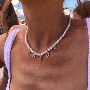May include: A pearl necklace with gold-colored letters spelling "BRIDE". The letters are embellished with small, clear crystals. The necklace is worn around the neck.