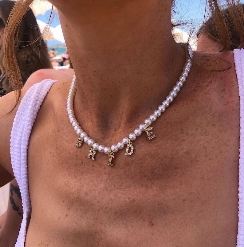 May include: A pearl necklace with gold-colored letters spelling "BRIDE". The letters are embellished with small, clear crystals. The necklace is worn around the neck.