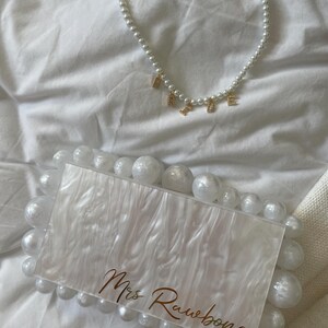 May include: A white pearl necklace with gold letter charms spelling "RACHEL" rests above a white and pearl-embellished clutch. The clutch features the text "Mrs Rawbone" in gold script, creating a cohesive bridal accessory set.