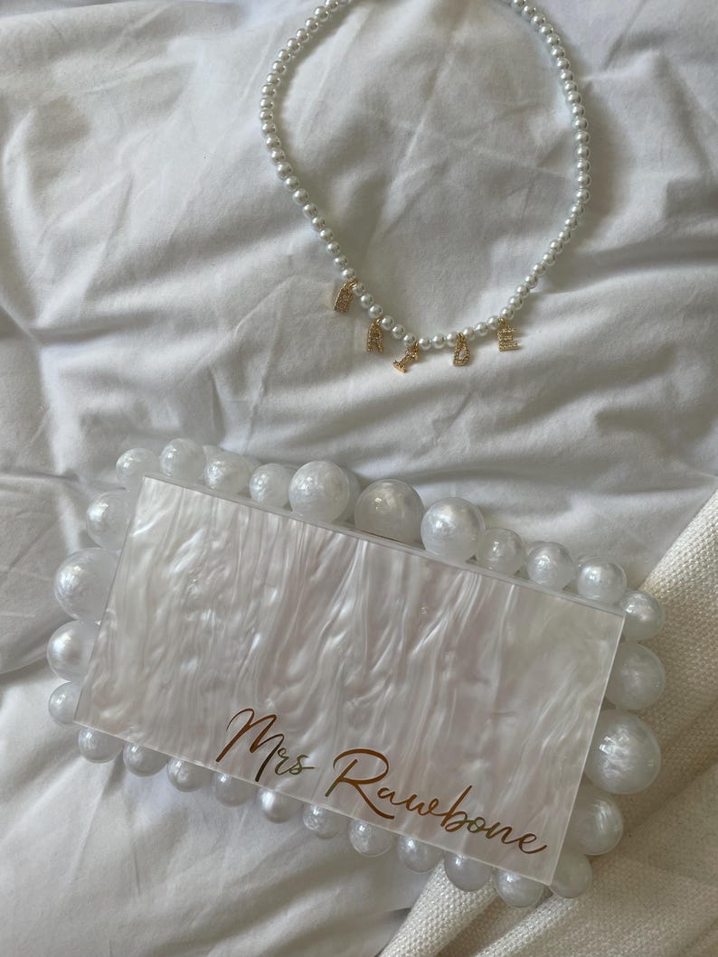 May include: A white pearl necklace with gold letter charms spelling "RACHEL" rests above a white and pearl-embellished clutch. The clutch features the text "Mrs Rawbone" in gold script, creating a cohesive bridal accessory set.
