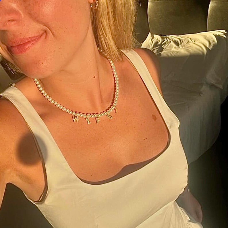 May include: A pearl necklace with gold-colored letters spelling "WIFE". The necklace is worn over a white dress. The image is taken in natural light.